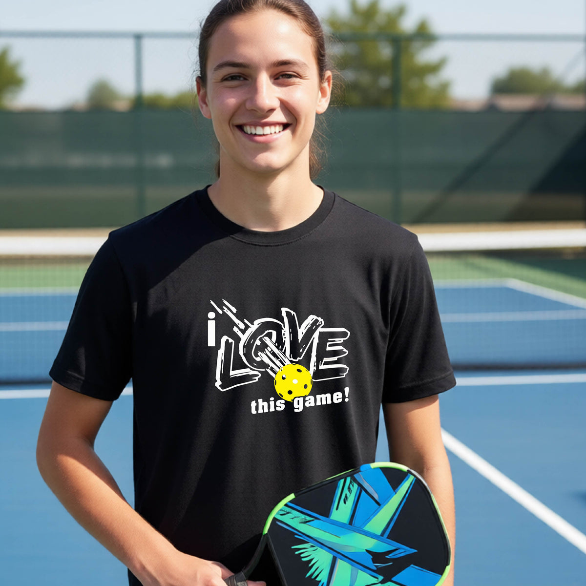 A young man laughing on a pickleball court wearing a black t-shirt with a white graphic that reads 'i LOVE this game!' and a yellow pickleball graphic.