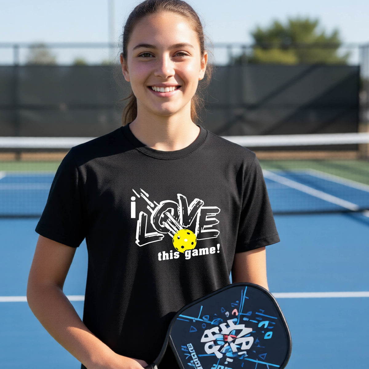 A young person laughing on a pickleball court wearing a black t-shirt with a white graphic that reads 'i LOVE this game!' and a yellow pickleball graphic.