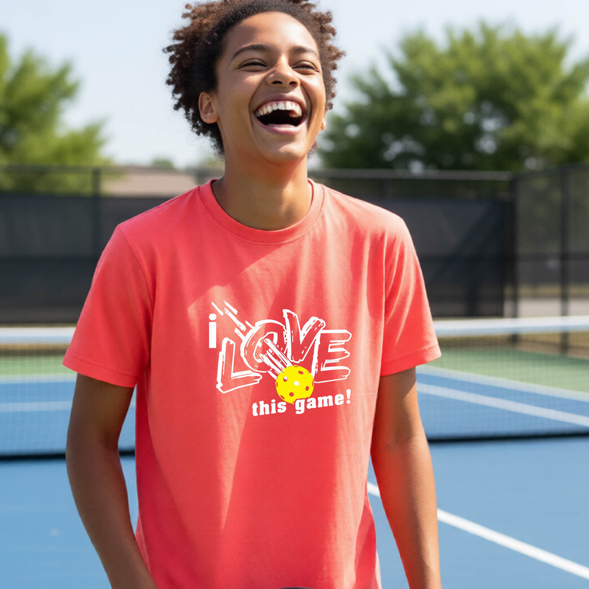 A young person laughing on a pickleball court wearing a coral pink t-shirt with a white graphic that reads 'i LOVE this game!' and a yellow pickleball graphic.