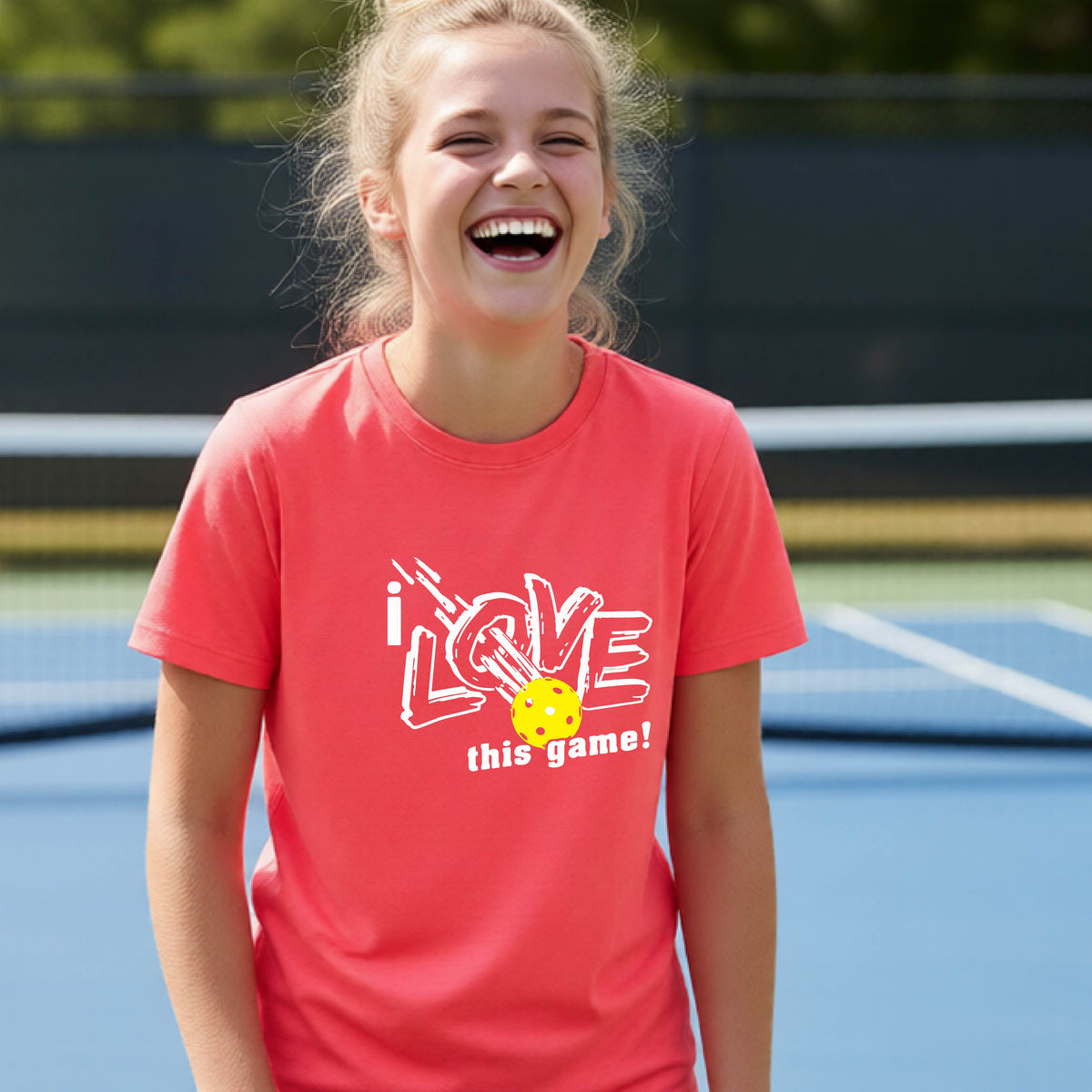 A young girl laughing on a pickleball court wearing a coral pink t-shirt with a white graphic that reads 'i LOVE this game!' and a yellow pickleball graphic.