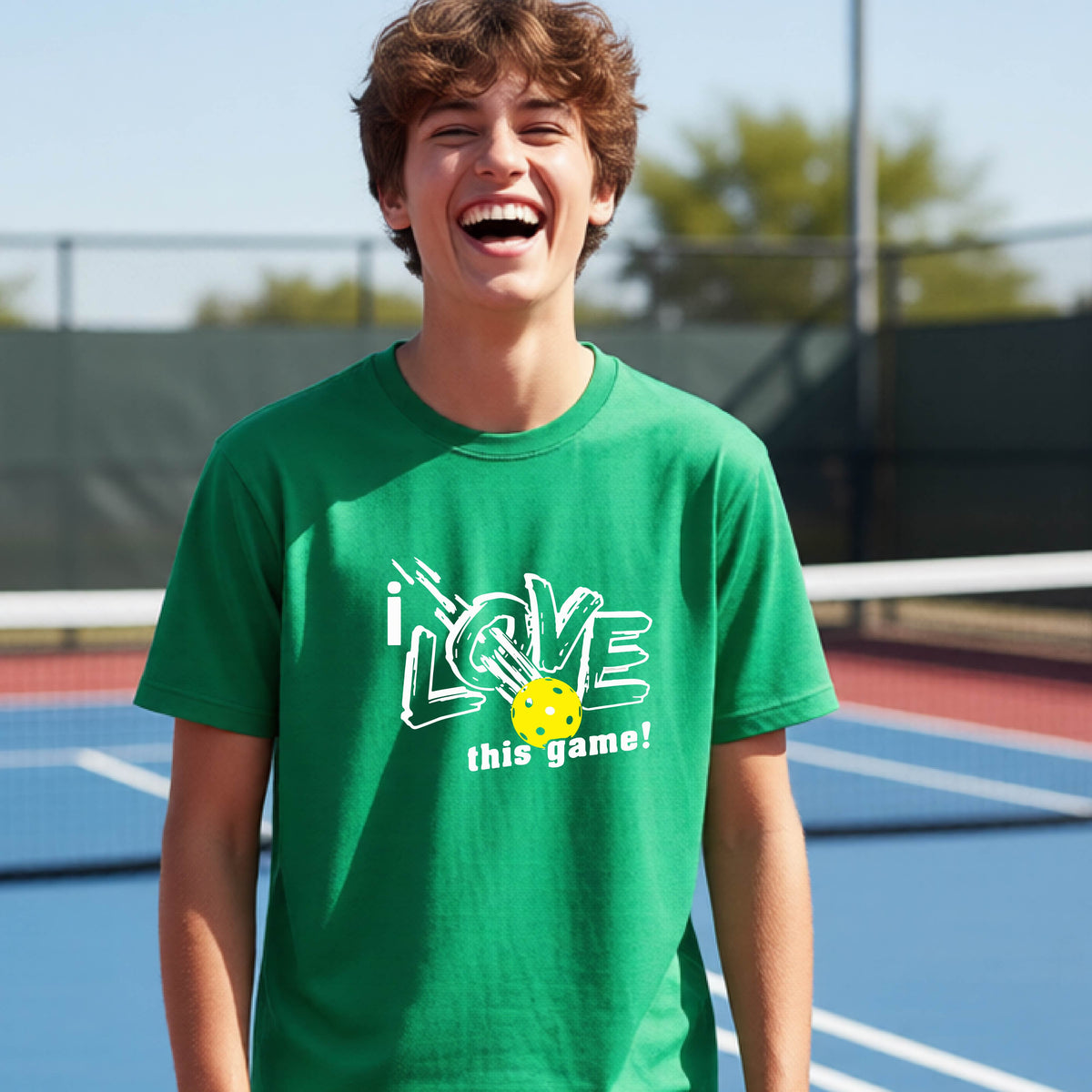 A young person laughing on a pickleball court wearing a green t-shirt with a white graphic that reads 'i LOVE this game!' and a yellow pickleball graphic.