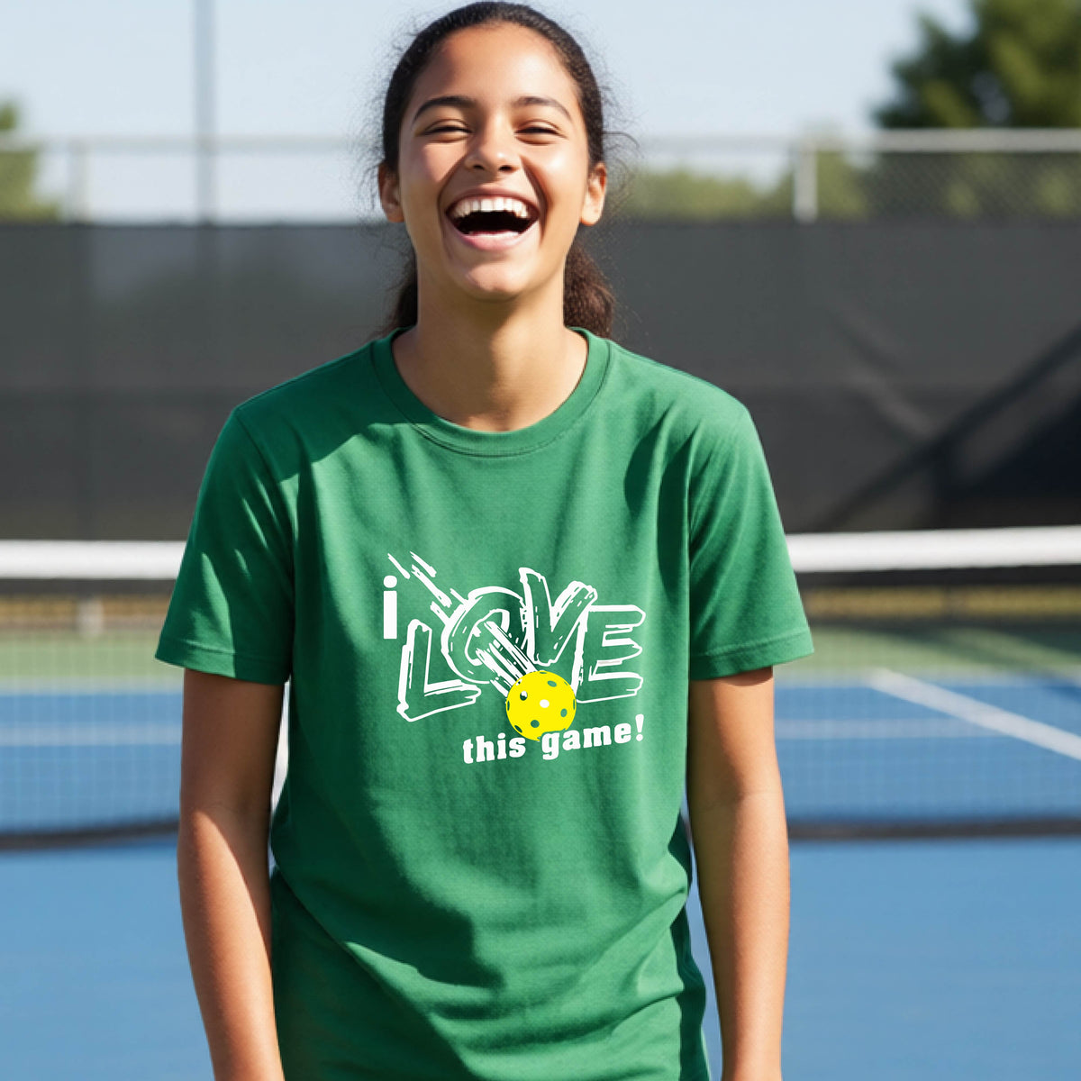 A young person laughing on a pickleball court wearing a green t-shirt with a white graphic that reads 'i LOVE this game!' and a yellow pickleball graphic.