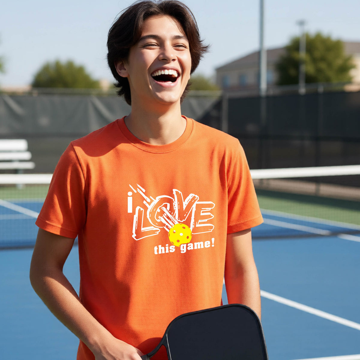 A young person laughing on a pickleball court wearing an orange t-shirt with a white graphic that reads 'i LOVE this game!' and a yellow pickleball graphic.