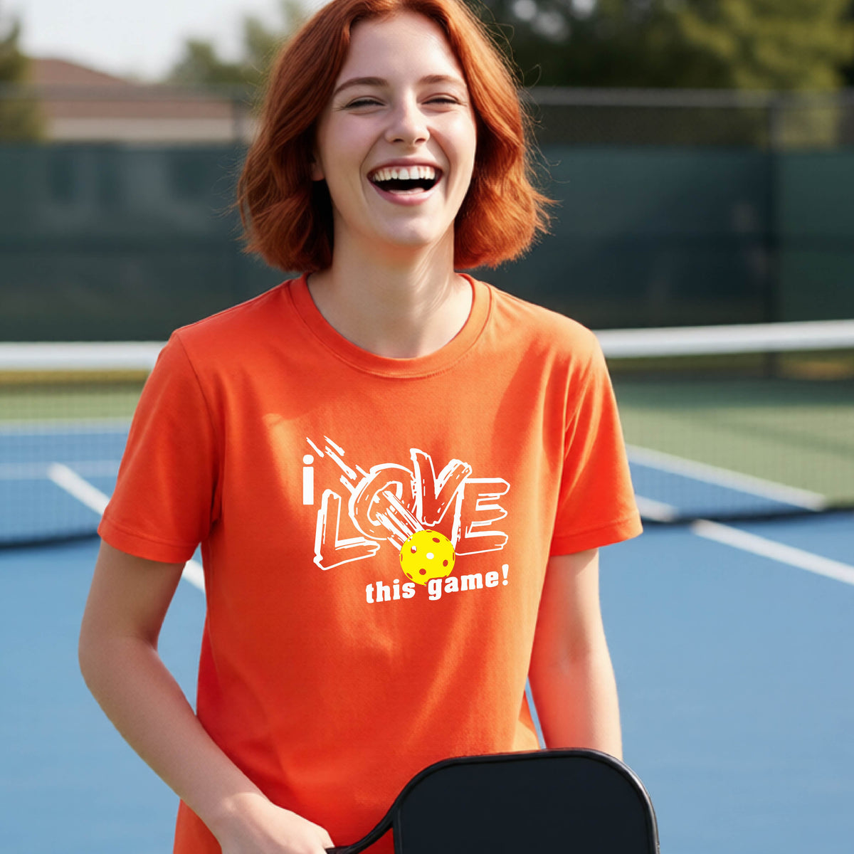 A young person laughing on a pickleball court wearing an orange t-shirt with a white graphic that reads 'i LOVE this game!' and a yellow pickleball graphic.