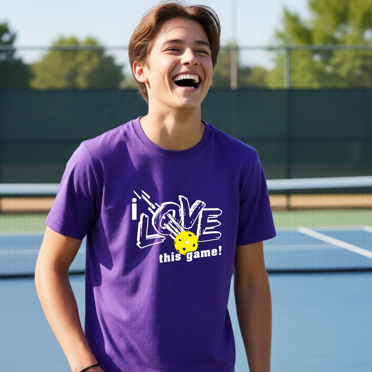 A young man laughing on a pickleball court wearing a purple t-shirt with a white graphic that reads 'i LOVE this game!' and a yellow pickleball graphic.