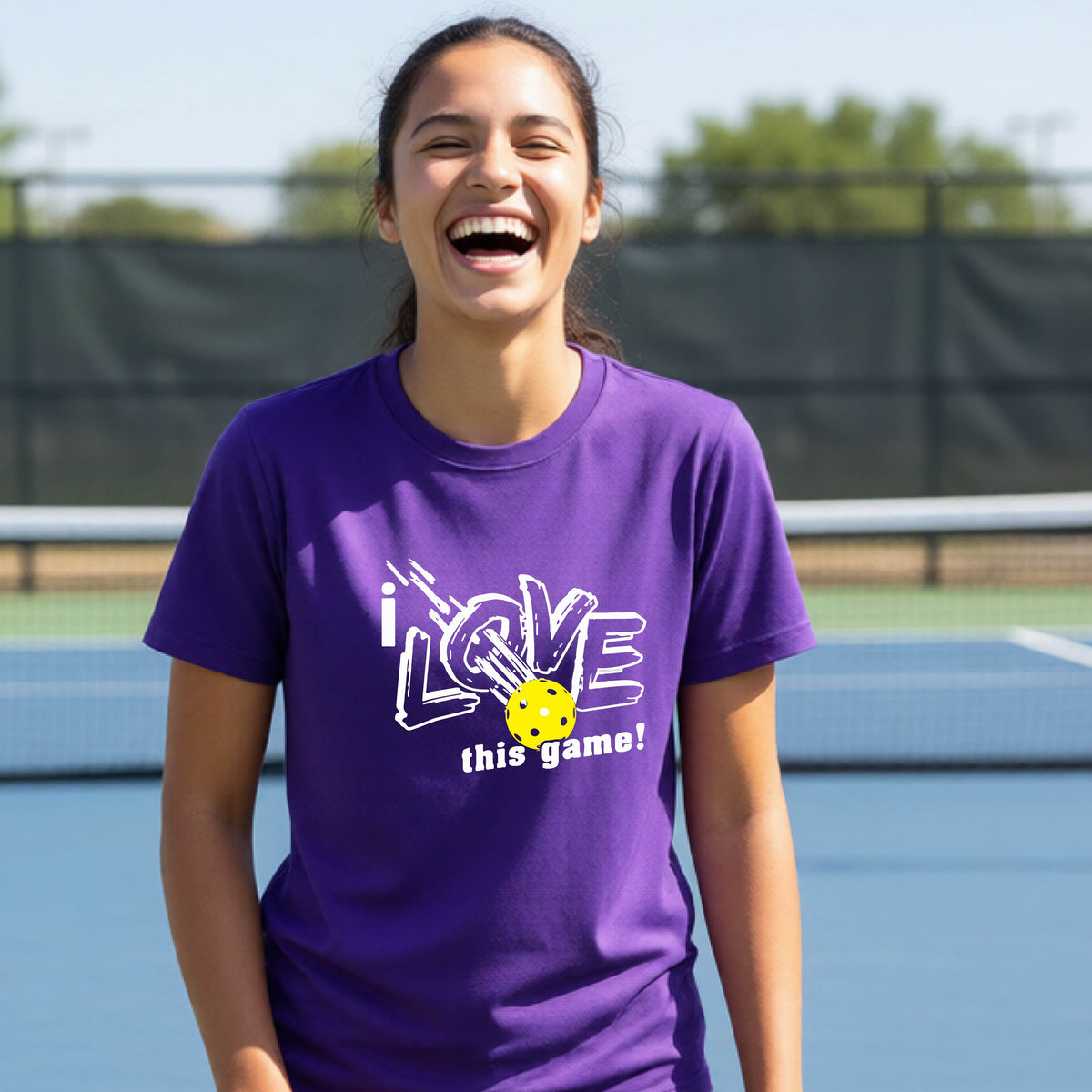 A young person laughing on a pickleball court wearing a purple t-shirt with a white graphic that reads 'i LOVE this game!' and a yellow pickleball graphic.