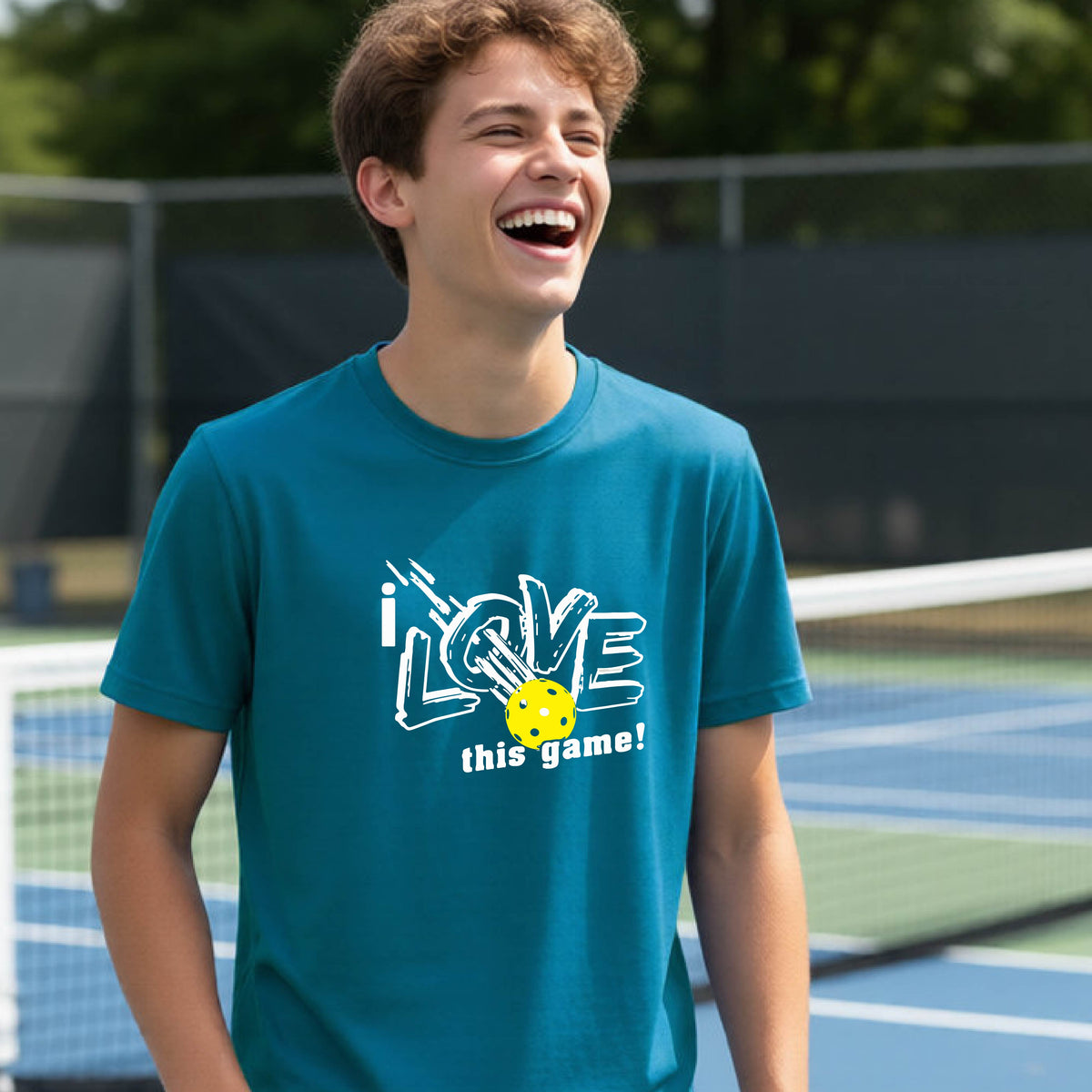A young person laughing on a pickleball court wearing a tropic blue t-shirt with a white graphic that reads 'i LOVE this game!' and a yellow pickleball graphic.