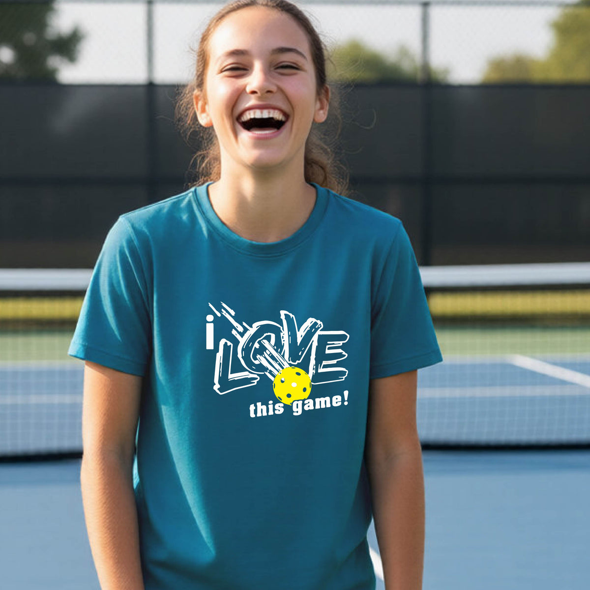 A young person laughing on a pickleball court wearing a teal blue t-shirt with a white graphic that reads 'i LOVE this game!' and a yellow pickleball graphic.