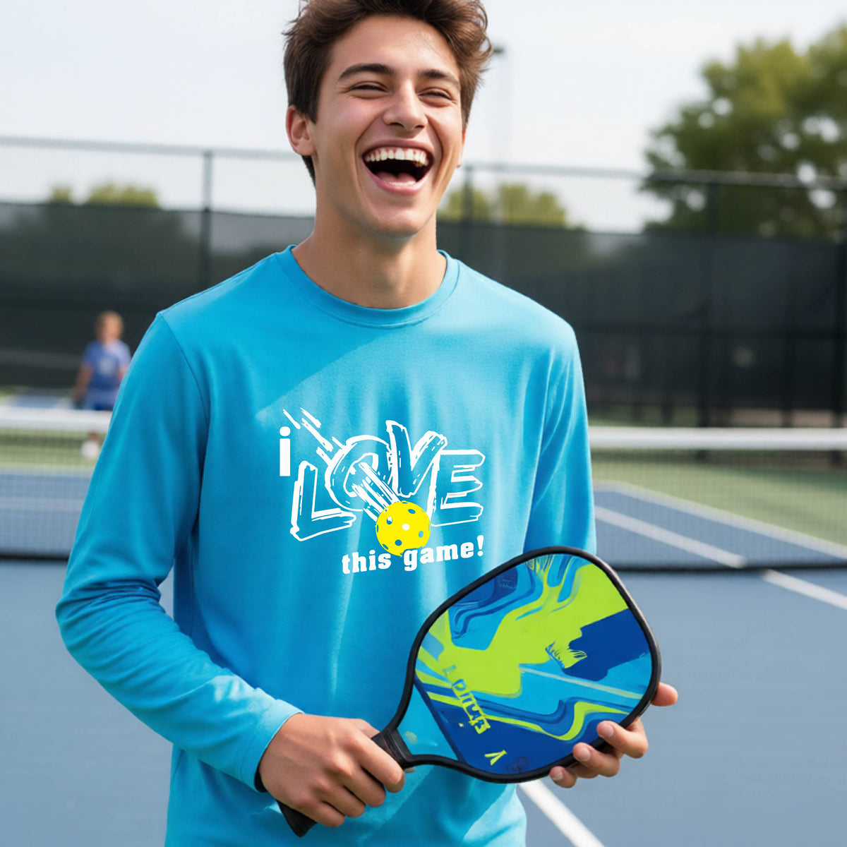 A young person laughing on a pickleball court wearing a bright blue long-sleeve t-shirt with a white graphic that reads 'i LOVE this game!' and a yellow pickleball illustration.