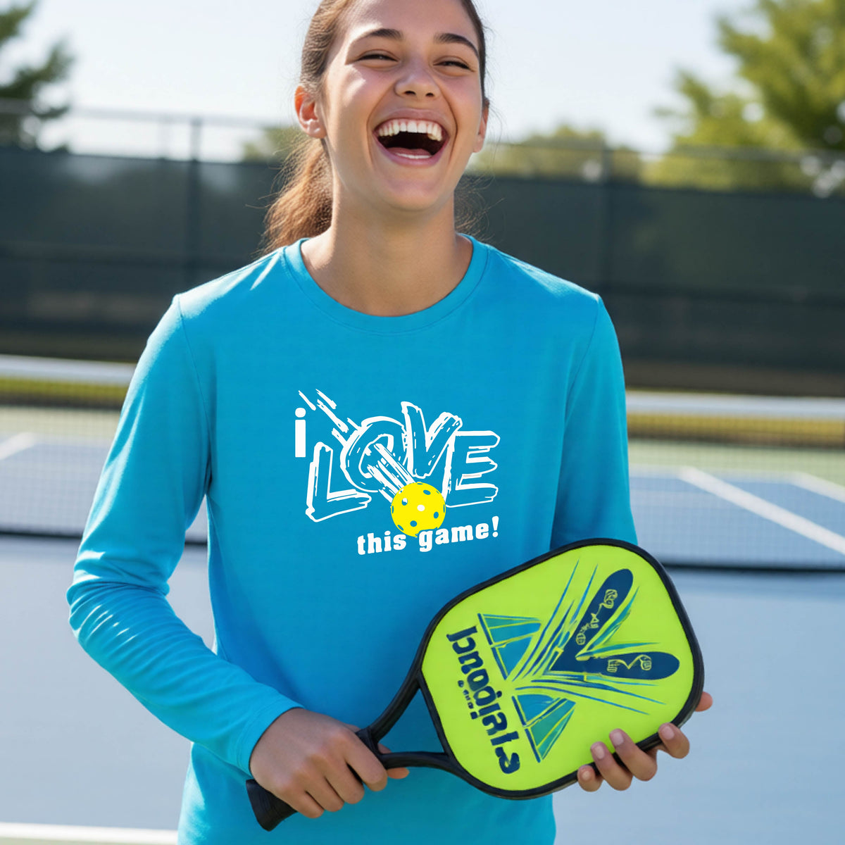 A young person laughing on a pickleball court wearing a bright blue long-sleeve t-shirt with a white graphic that reads 'i LOVE this game!' and a yellow pickleball illustration.