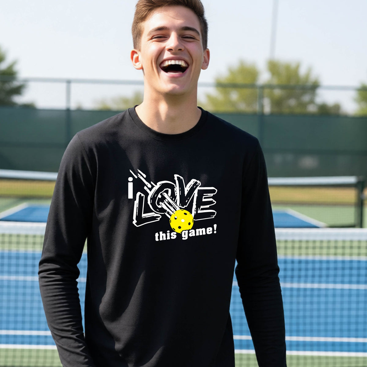 A young person laughing on a pickleball court wearing a black long-sleeve t-shirt with a white graphic that reads 'i LOVE this game!' and a yellow pickleball illustration.