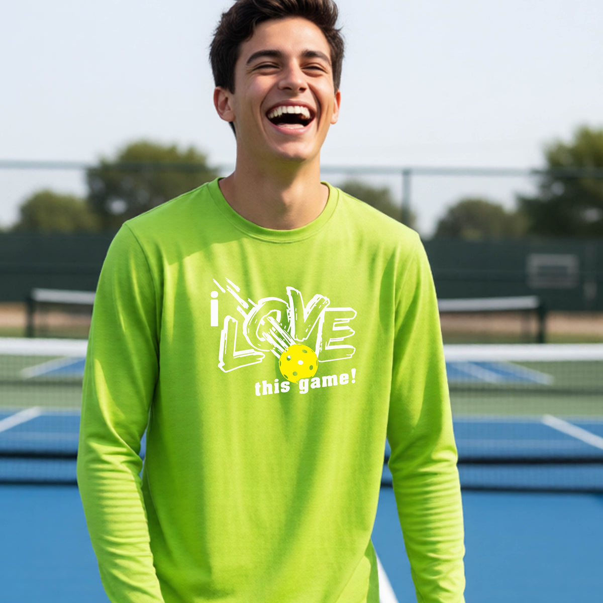 A young person laughing on a pickleball court wearing a lime green long-sleeve t-shirt with a white graphic that reads 'i LOVE this game!' and a yellow pickleball illustration.