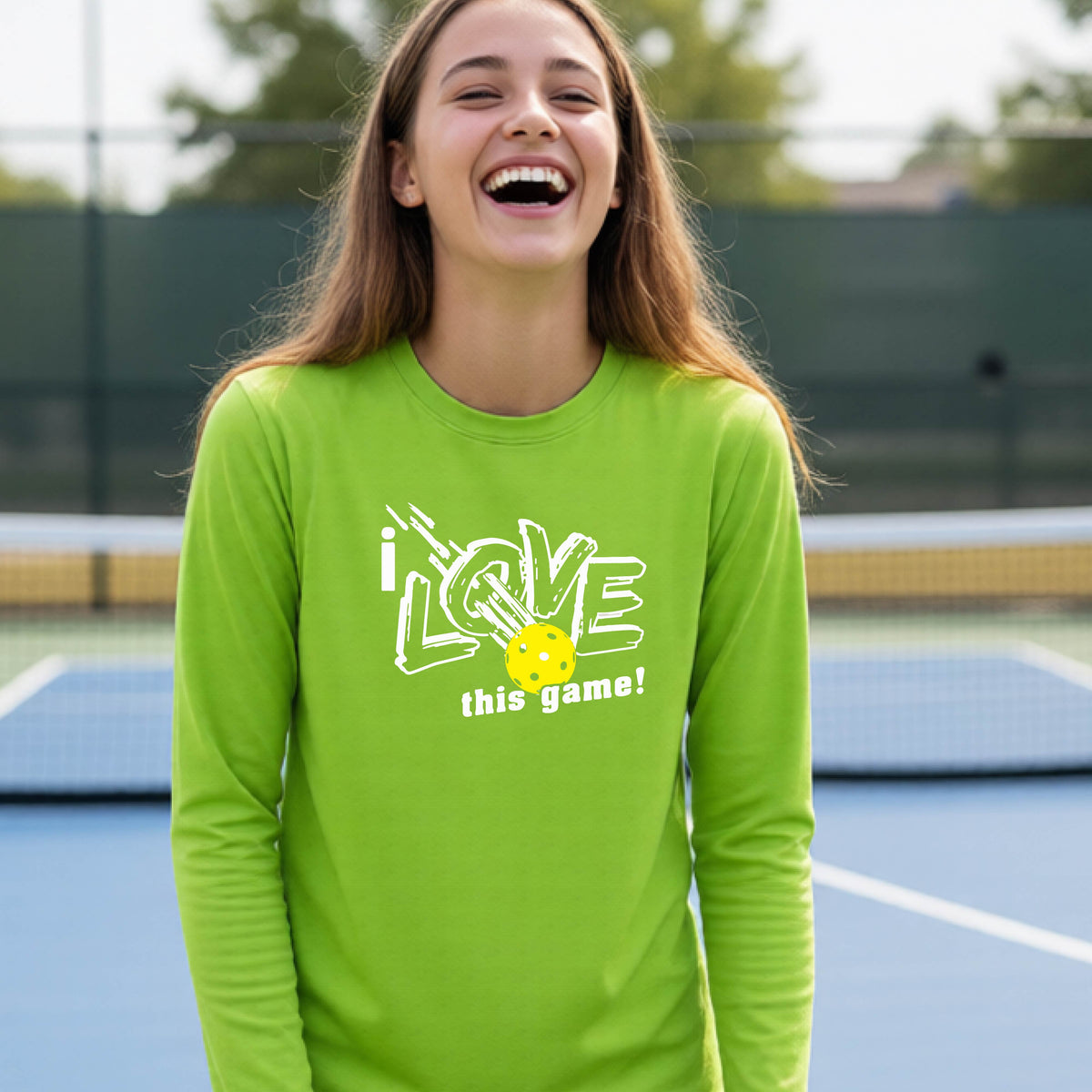 A young person laughing on a pickleball court wearing a lime green long-sleeve t-shirt with a white graphic that reads 'i LOVE this game!' and a yellow pickleball illustration.