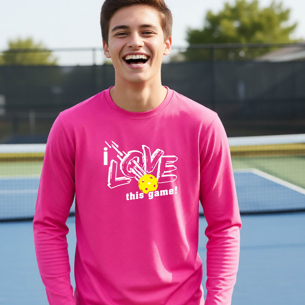 A young person laughing on a pickleball court wearing a pink long-sleeve t-shirt with a white graphic that reads 'i LOVE this game!' and a yellow pickleball illustration.