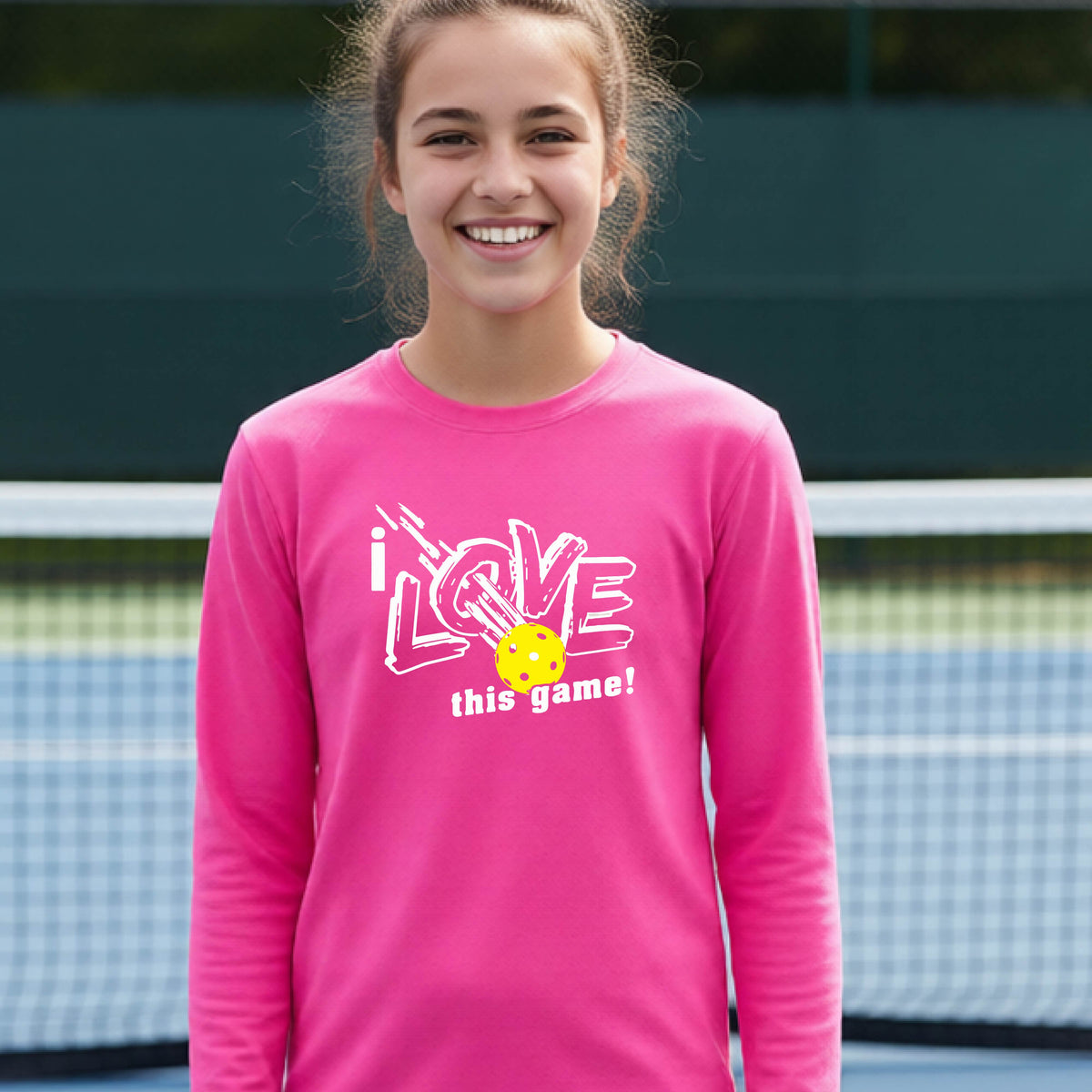 A young person laughing on a pickleball court wearing a pink long-sleeve t-shirt with a white graphic that reads 'i LOVE this game!' and a yellow pickleball illustration.