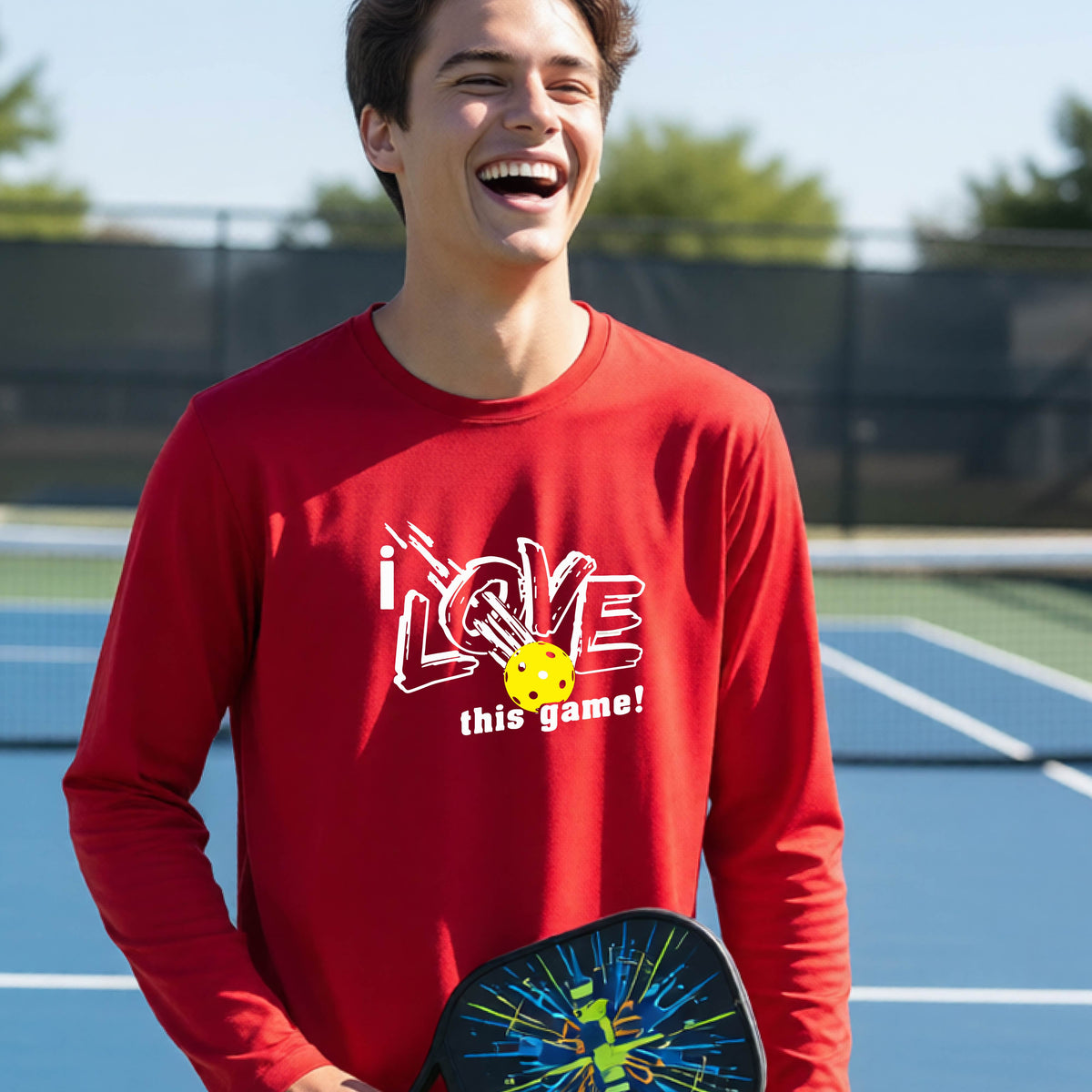 A young person laughing on a pickleball court wearing a red long-sleeve t-shirt with a white graphic that reads 'i LOVE this game!' and a yellow pickleball illustration.