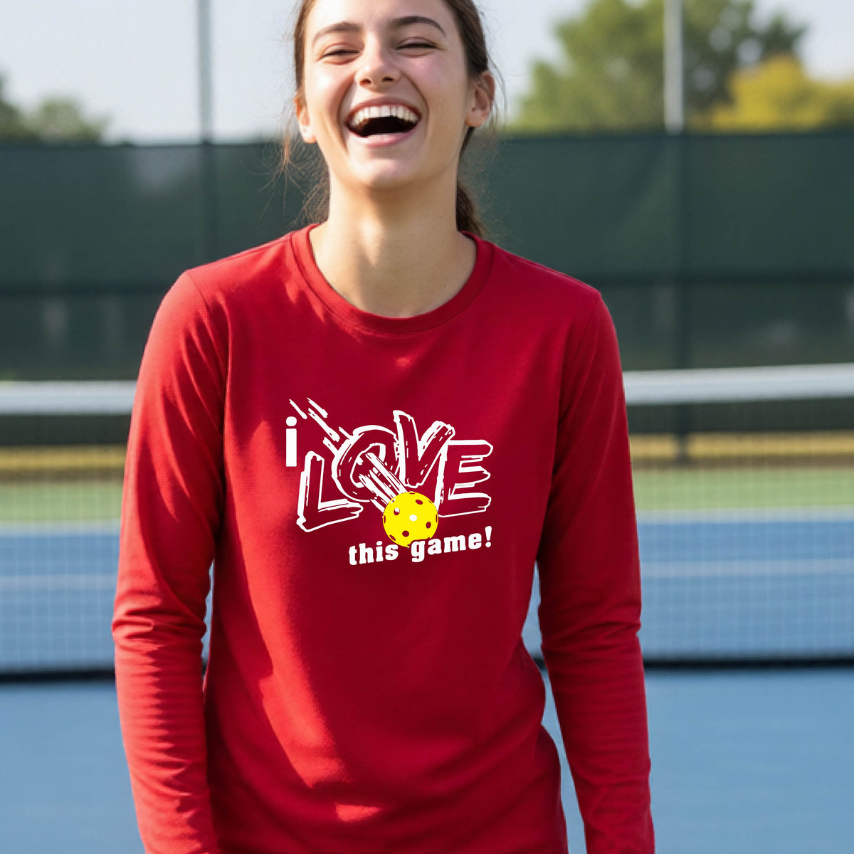 A young person laughing on a pickleball court wearing a red long-sleeve t-shirt with a white graphic that reads 'i LOVE this game!' and a yellow pickleball illustration.