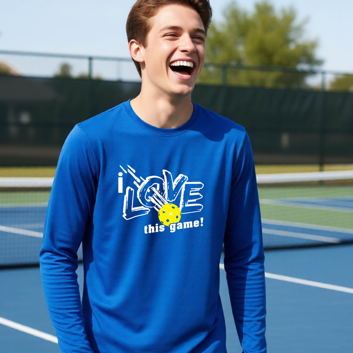 A young person laughing on a pickleball court wearing a royal blue long-sleeve t-shirt with a white graphic that reads 'i LOVE this game!' and a yellow pickleball illustration.