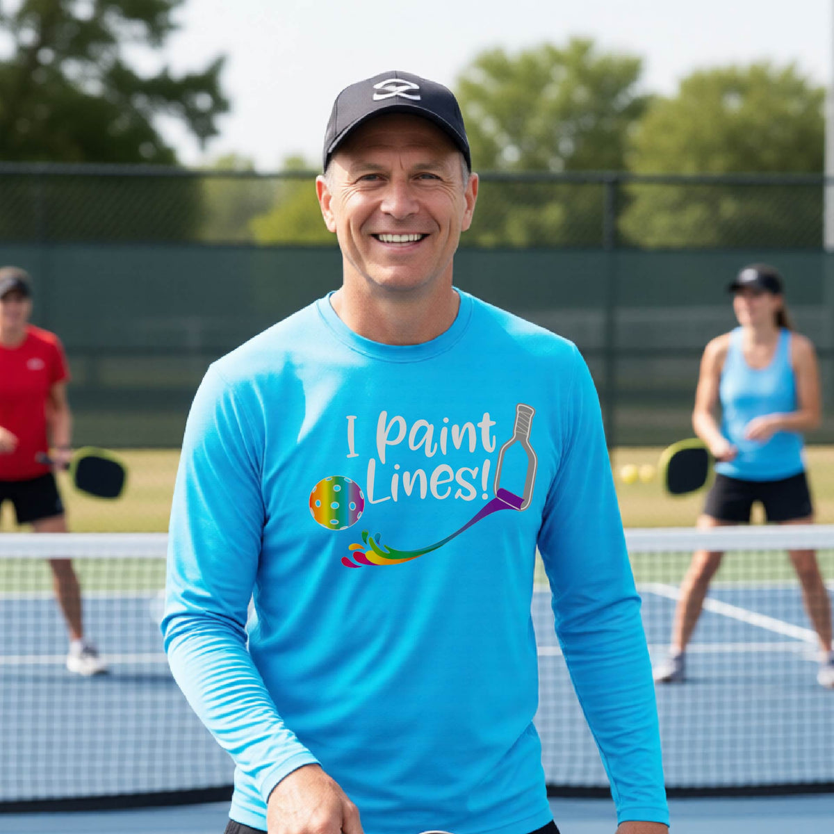 A close-up of a man wearing a bright blue long-sleeve t-shirt with a graphic on the front that has white text "I PAINT LINES'  with a white pickleball paddle painting multi-colored lines leading a mutli-colored pickleball.