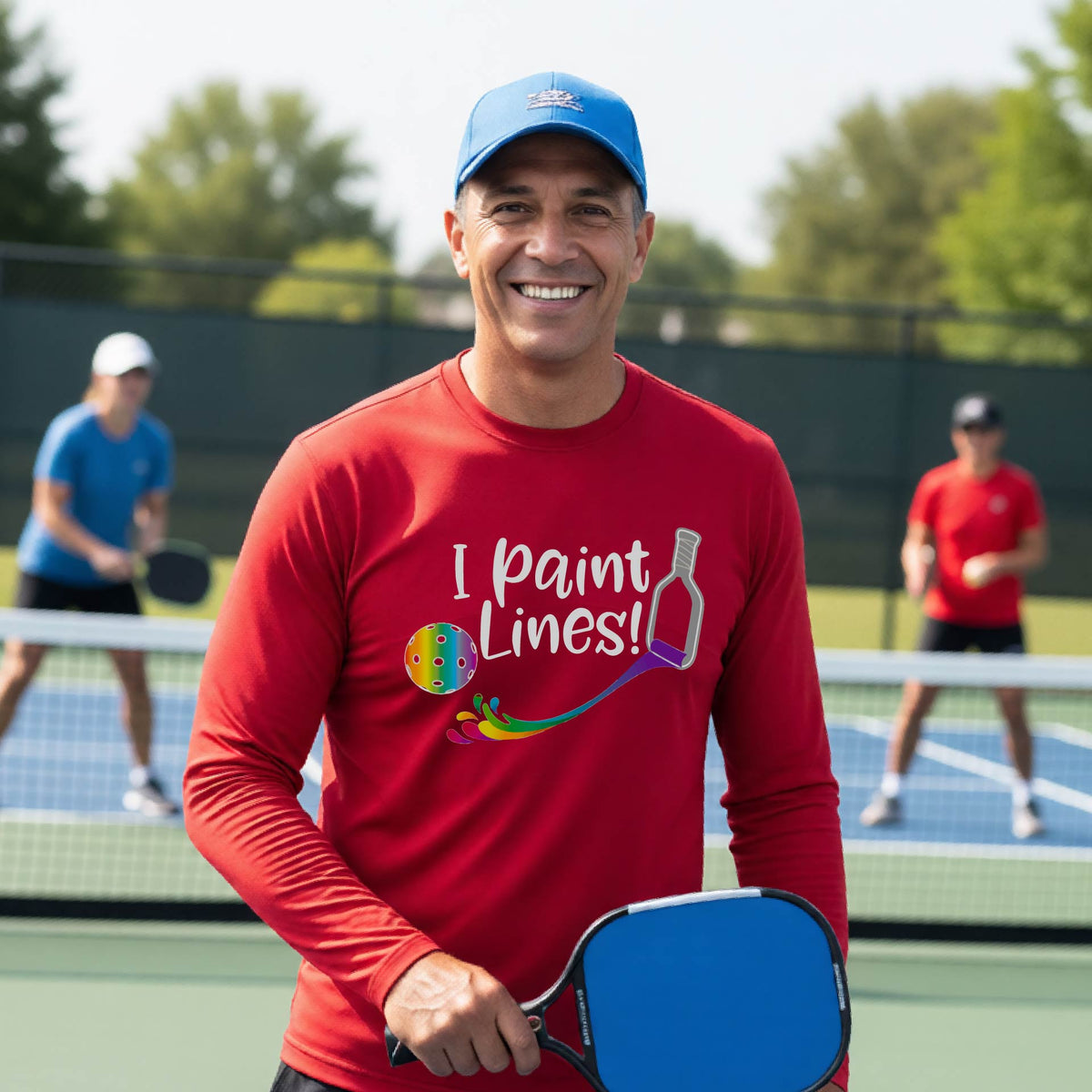 A close-up of a man wearing a red long-sleeve t-shirt with a graphic on the front that has white text "I PAINT LINES'  with a white pickleball paddle painting multi-colored lines leading a mutli-colored pickleball.