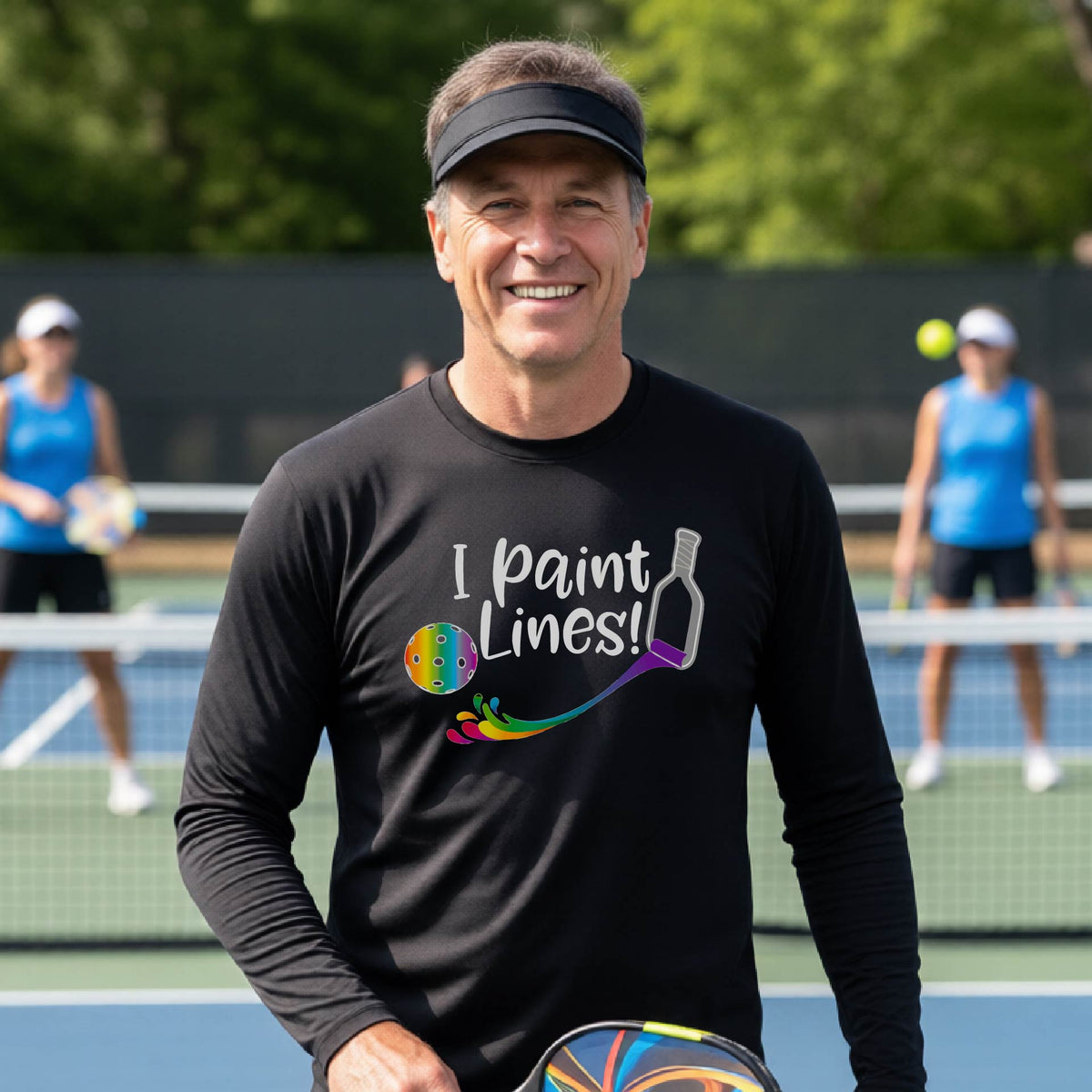 A close-up of a man wearing a black long-sleeve t-shirt with a graphic on the front that has white text "I PAINT LINES'  with a white pickleball paddle painting multi-colored lines leading a mutli-colored pickleball.