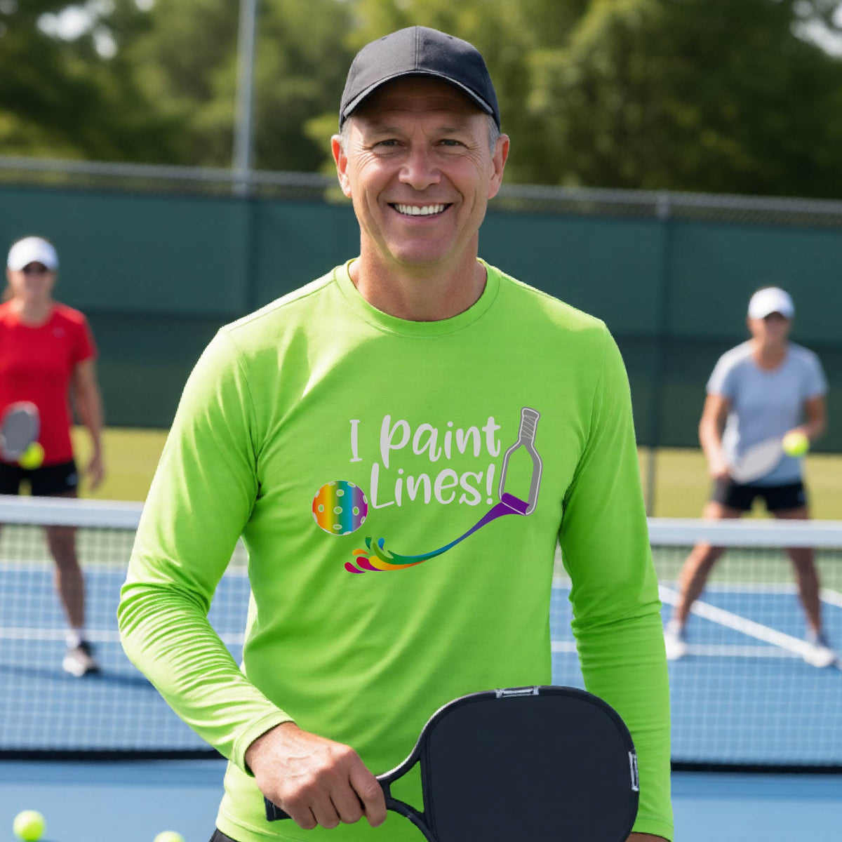 A close-up of a man wearing a lime green long-sleeve t-shirt with a graphic on the front that has white text "I PAINT LINES'  with a white pickleball paddle painting multi-colored lines leading a mutli-colored pickleball.