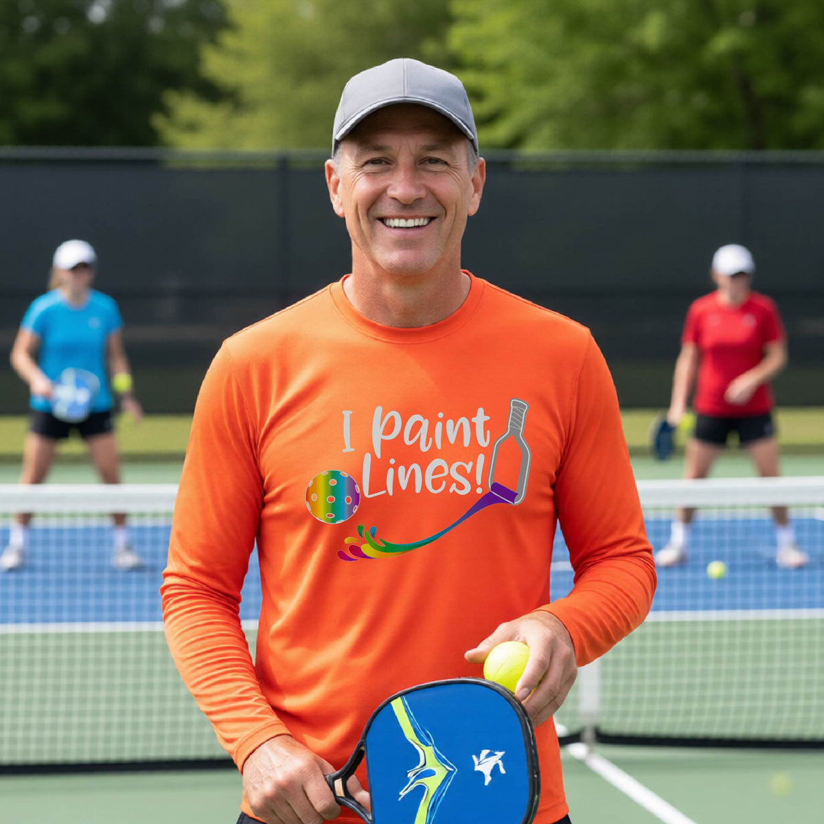 A close-up of a man wearing an orange long-sleeve t-shirt with a graphic on the front that has white text "I PAINT LINES'  with a white pickleball paddle painting multi-colored lines leading a mutli-colored pickleball.