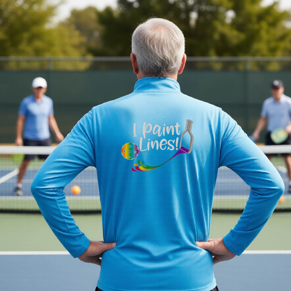A man on a pickleball court seen from behind, wearing a bright blue long-sleeve shirt with a colorful 'I paint Lines!' graphic on the back including a pickleball paddle painting mutli-colored lines going to a multi-colored pickleball