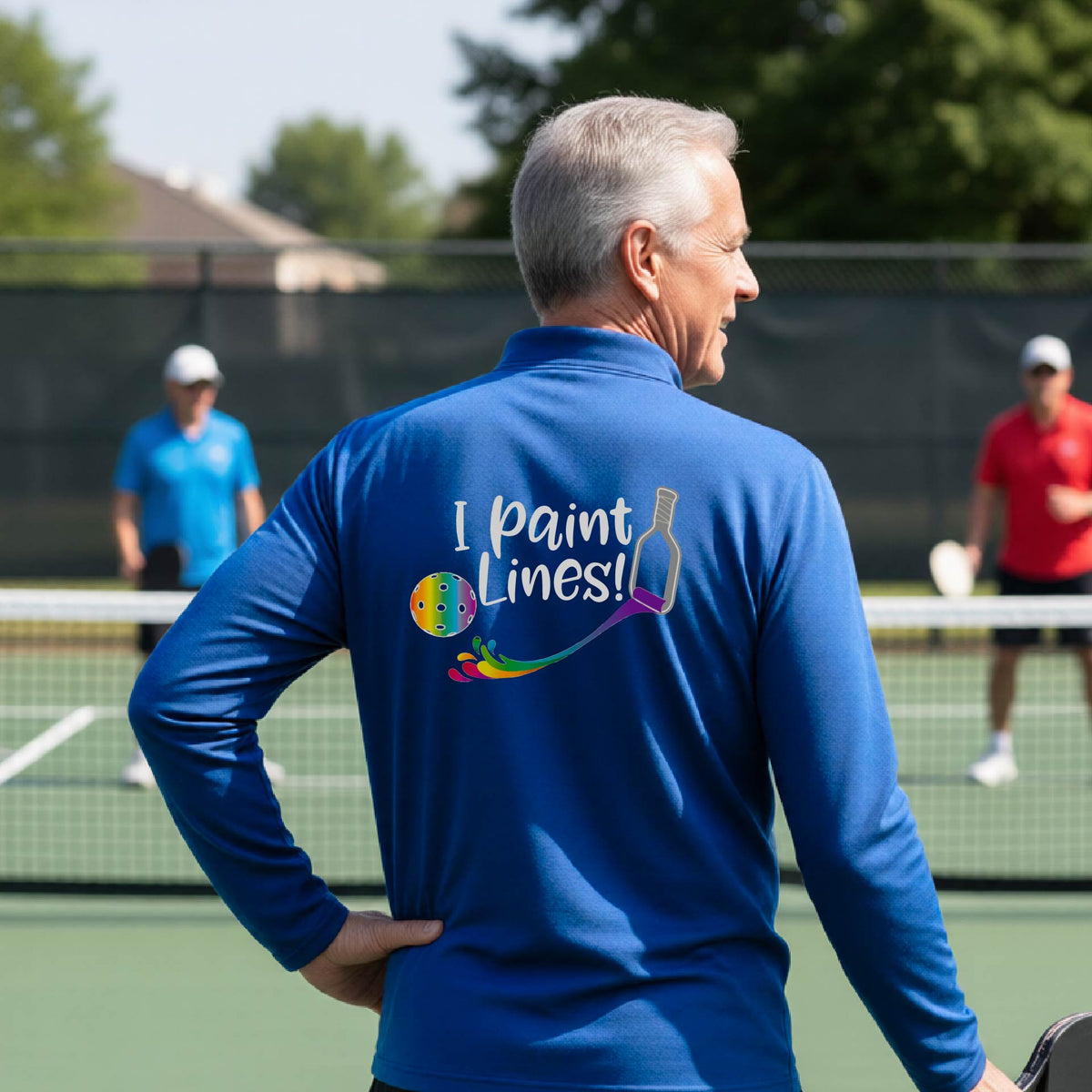 A man on a pickleball court seen from behind, wearing a bright royal blue long-sleeve shirt with a colorful 'I paint Lines!' graphic on the back including a pickleball paddle painting mutli-colored lines going to a multi-colored pickleball