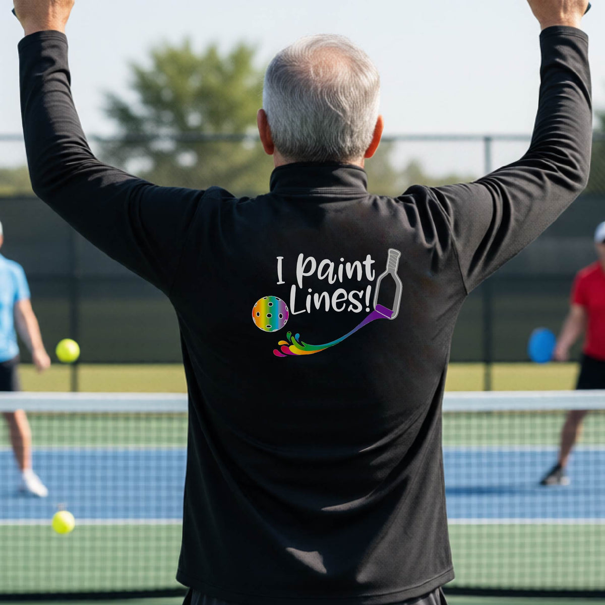 A man on a pickleball court seen from behind, wearing a black long-sleeve shirt with a colorful 'I paint Lines!' graphic on the back including a pickleball paddle painting mutli-colored lines going to a multi-colored pickleball