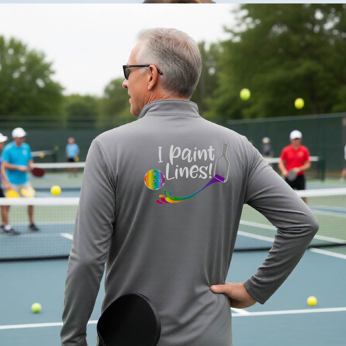 A man on a pickleball court seen from behind, wearing a gray long-sleeve shirt with a colorful 'I paint Lines!' graphic on the back including a pickleball paddle painting mutli-colored lines going to a multi-colored pickleball