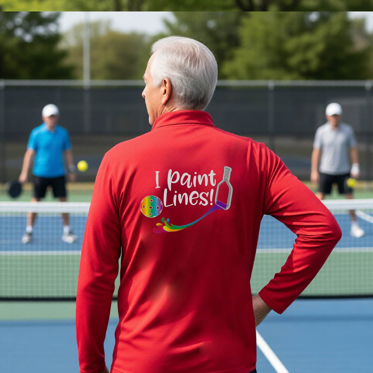 A man on a pickleball court seen from behind, wearing a bright red long-sleeve shirt with a colorful 'I paint Lines!' graphic on the back including a pickleball paddle painting mutli-colored lines going to a multi-colored pickleball