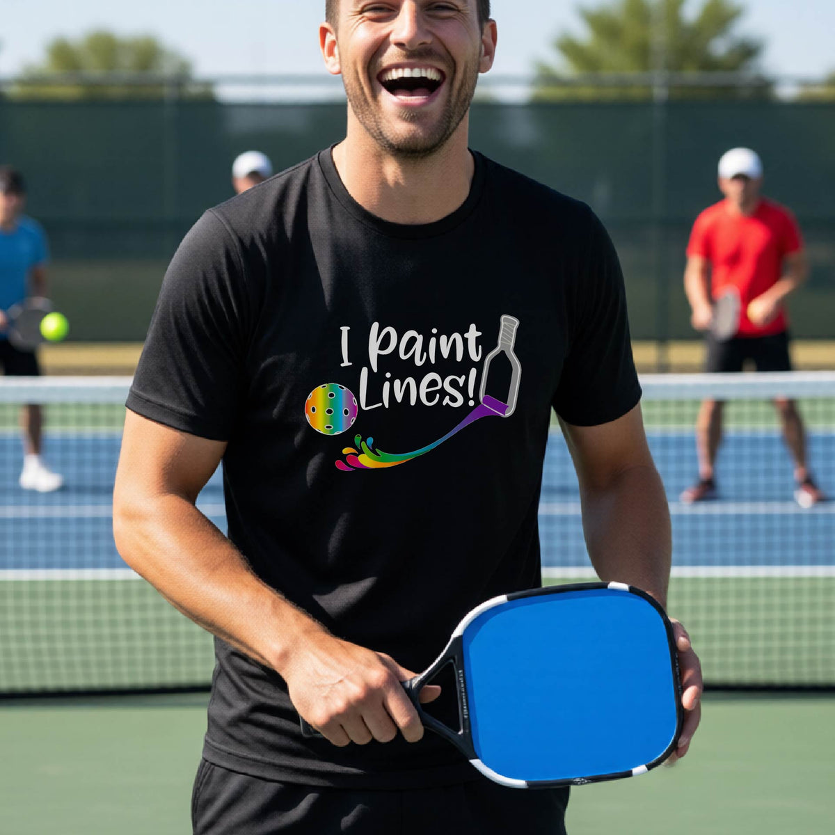 A close-up of a woman wearing a black short-sleeve t-shirt with a graphic on the front that has white text "I PAINT LINES'  with a white pickleball paddle painting multi-colored lines leading a mutli-colored pickleball.