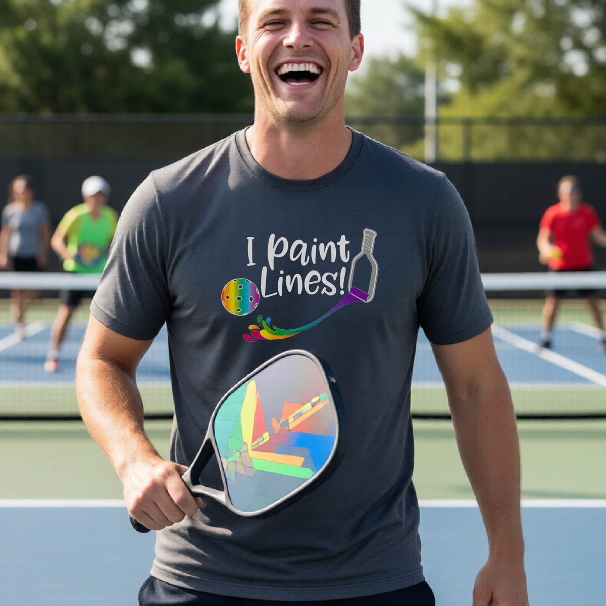 A close-up of a woman wearing a gray short-sleeve t-shirt with a graphic on the front that has white text "I PAINT LINES'  with a white pickleball paddle painting multi-colored lines leading a mutli-colored pickleball.
