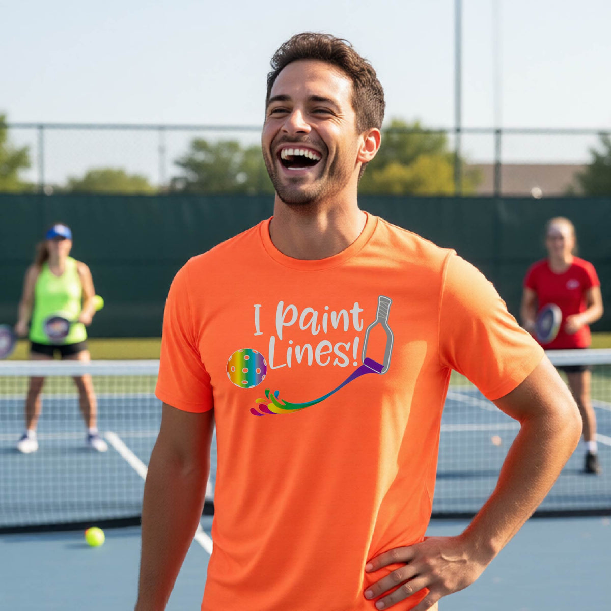 A close-up of a woman wearing an orange short-sleeve t-shirt with a graphic on the front that has white text "I PAINT LINES'  with a white pickleball paddle painting multi-colored lines leading a mutli-colored pickleball.