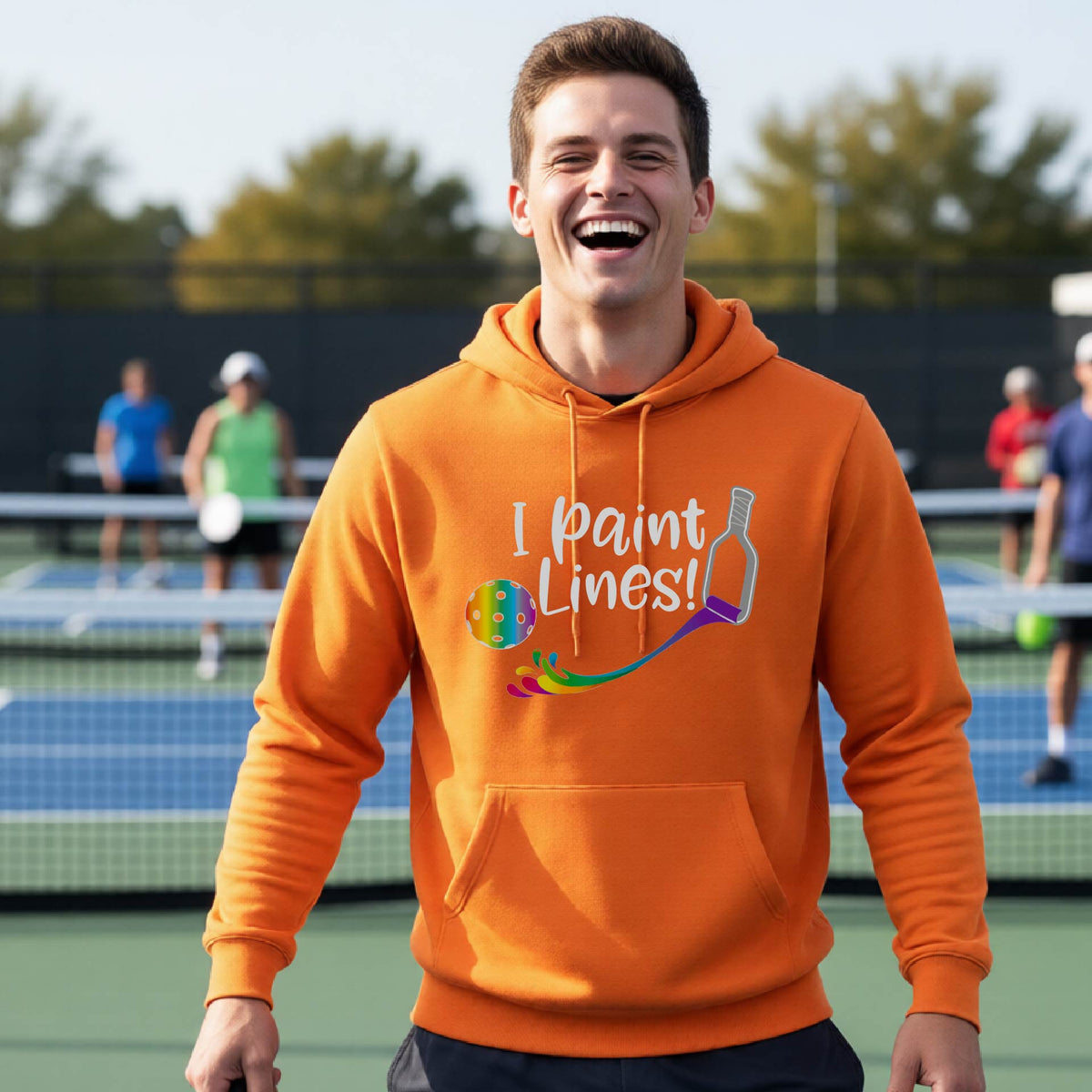 A man on an outdoor pickleball court wearing an orange unisex hoodie pullover with a graphic that reads 'THIS IS MY PICKLEBALL SHIRT' in white with a yellow pickleball graphic on the back.