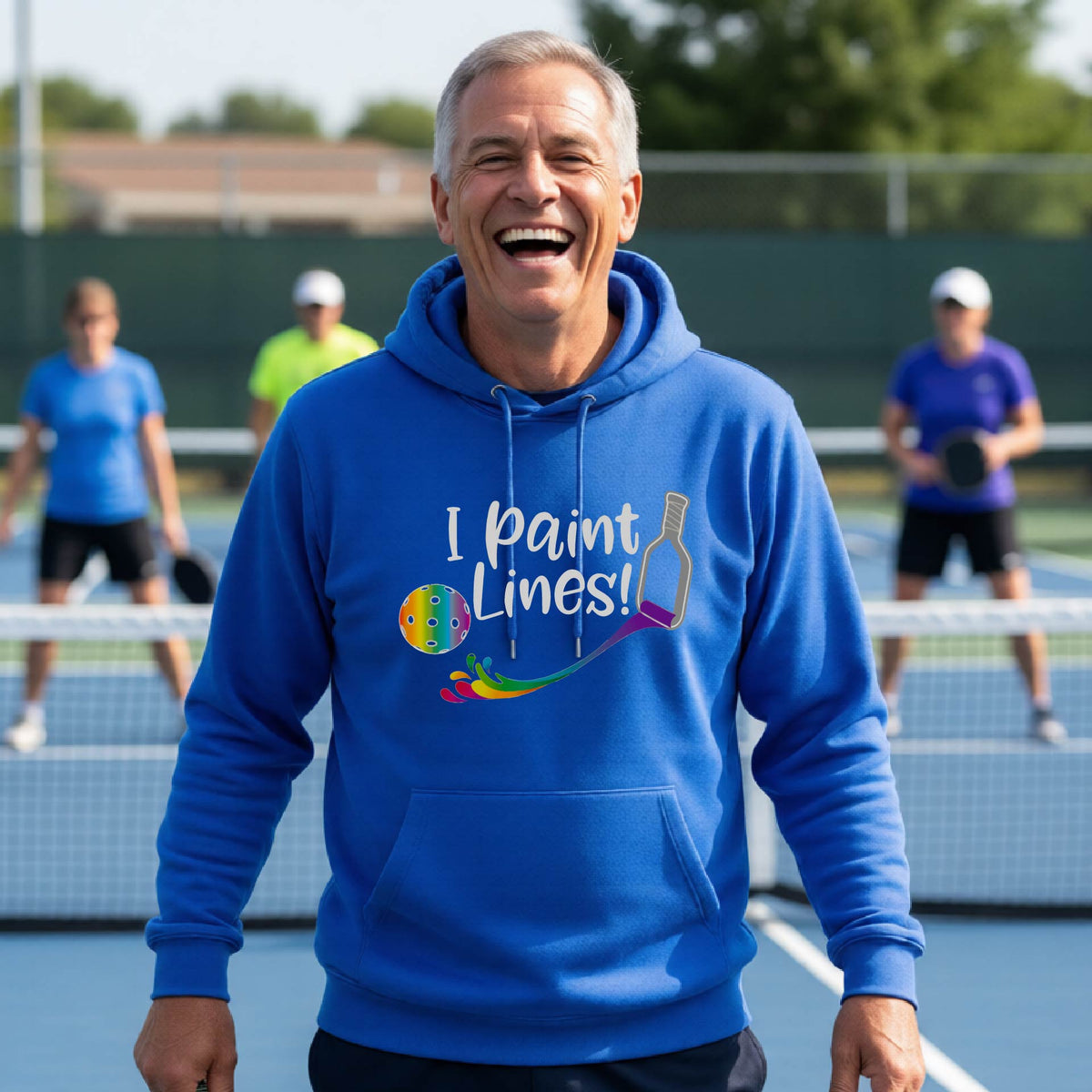 A man on an outdoor pickleball court wearing a royal blue unisex hoodie pullover with a graphic that reads 'THIS IS MY PICKLEBALL SHIRT' in white with a yellow pickleball graphic on the back.