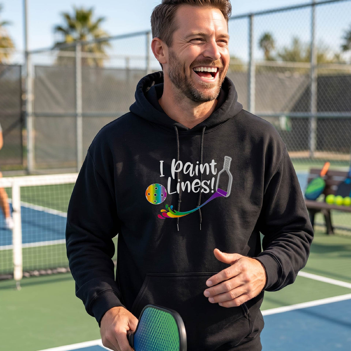 A man on an outdoor pickleball court wearing a black unisex hoodie pullover with a graphic that reads 'THIS IS MY PICKLEBALL SHIRT' in white with a yellow pickleball graphic on the back.