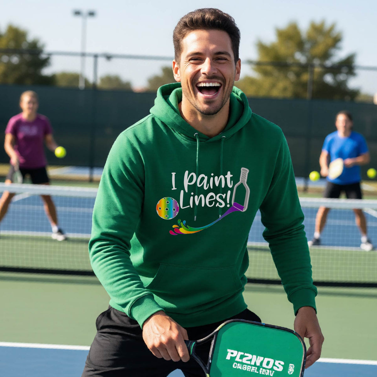 A man on an outdoor pickleball court wearing a green unisex hoodie pullover with a graphic that reads 'THIS IS MY PICKLEBALL SHIRT' in white with a yellow pickleball graphic on the back.