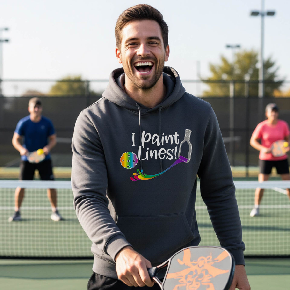 A man on an outdoor pickleball court wearing a gray unisex hoodie pullover with a graphic that reads 'THIS IS MY PICKLEBALL SHIRT' in white with a yellow pickleball graphic on the back.