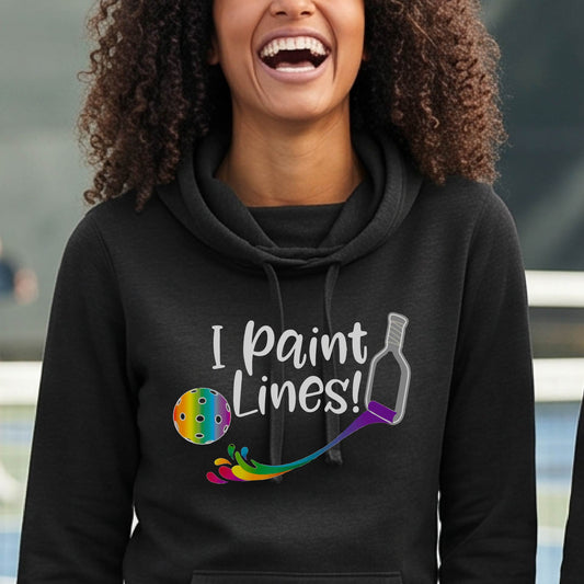 A close-up of a woman wearing a black long-sleeve cowl-neck hoodie pullover with a graphic on the front that has white text "I PAINT LINES'  with a white pickleball paddle painting multi-colored lines leading a mutli-colored pickleball.