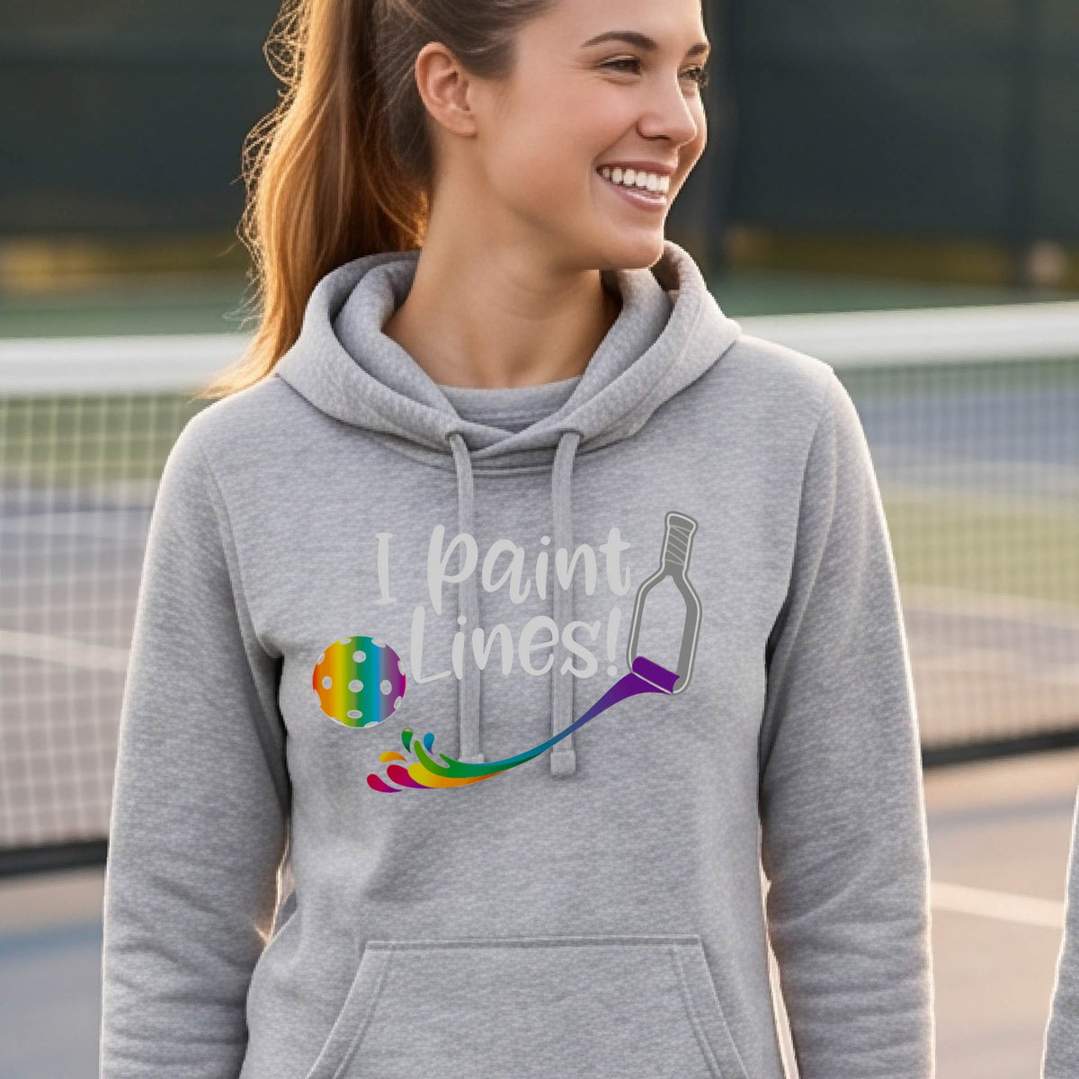 A close-up of a woman wearing a gray long-sleeve cowl-neck hoodie pullover with a graphic on the front that has white text "I PAINT LINES'  with a white pickleball paddle painting multi-colored lines leading a mutli-colored pickleball.