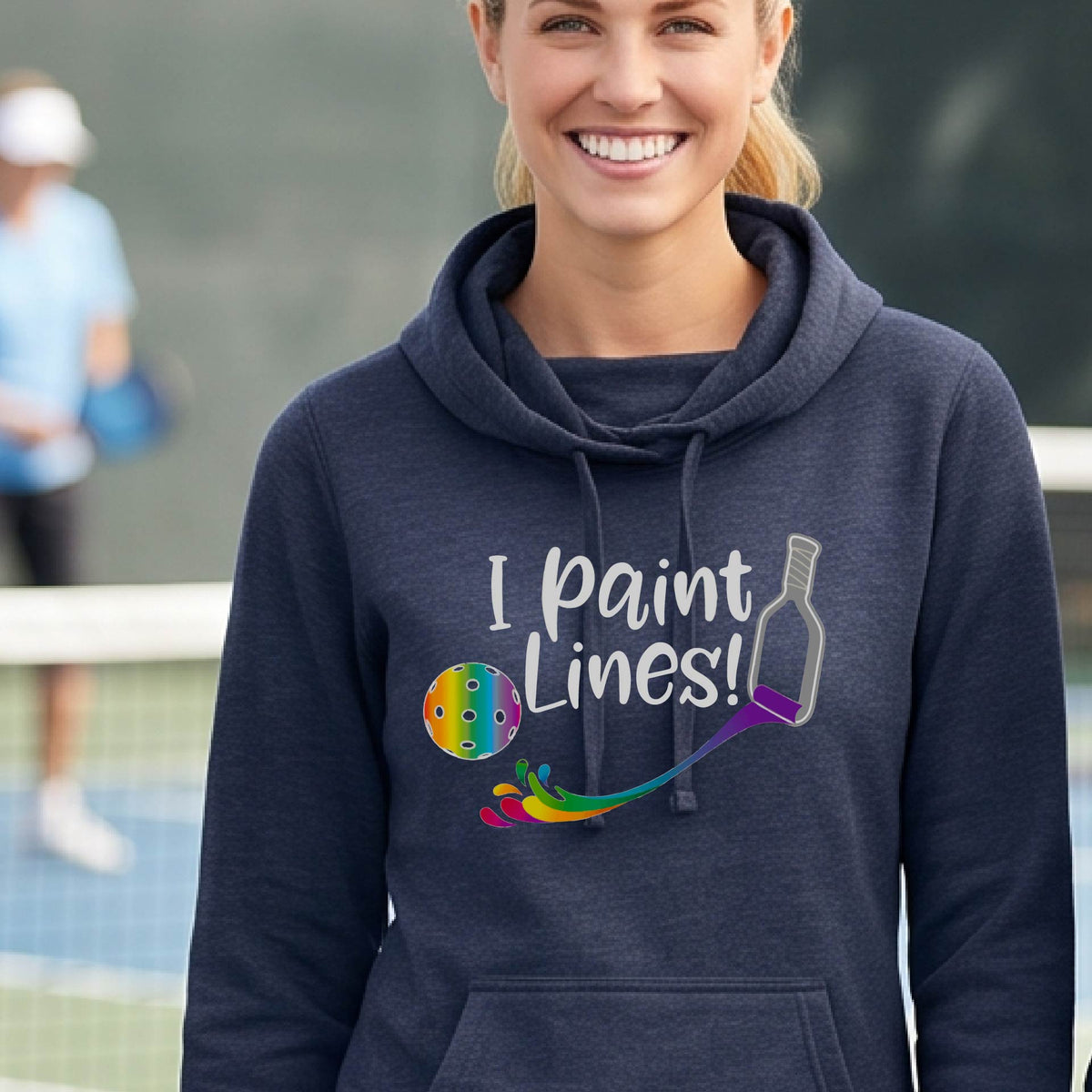 A close-up of a woman wearing a navy blue long-sleeve cowl-neck hoodie pullover with a graphic on the front that has white text "I PAINT LINES'  with a white pickleball paddle painting multi-colored lines leading a mutli-colored pickleball.