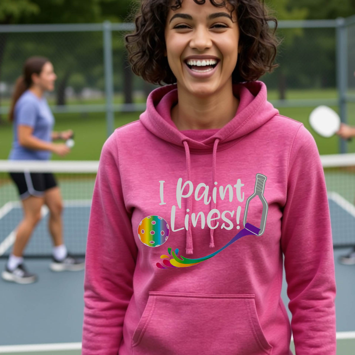 A close-up of a woman wearing a pink long-sleeve cowl-neck hoodie pullover with a graphic on the front that has white text "I PAINT LINES'  with a white pickleball paddle painting multi-colored lines leading a mutli-colored pickleball.