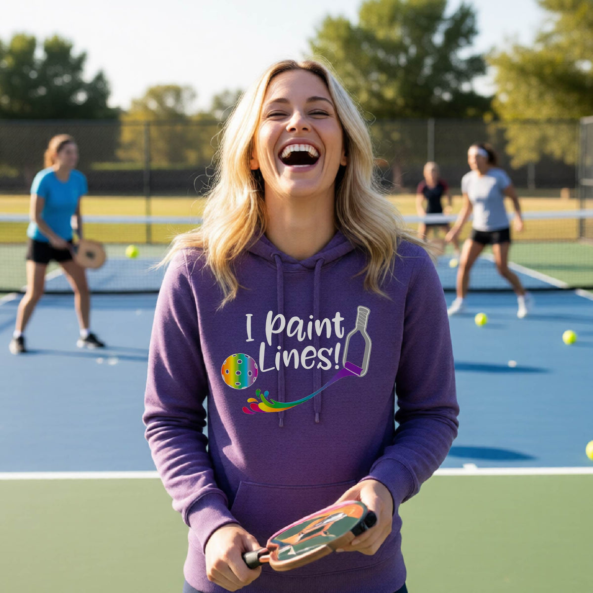 A woman wearing a purple fitted hoodie pullover with a graphic on the front that has white text "I PAINT LINES'  with a white pickleball paddle painting multi-colored lines leading a mutli-colored pickleball.