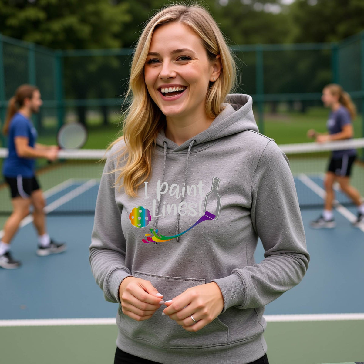 A woman wearing a gray fitted hoodie pullover with a graphic on the front that has white text "I PAINT LINES'  with a white pickleball paddle painting multi-colored lines leading a mutli-colored pickleball.