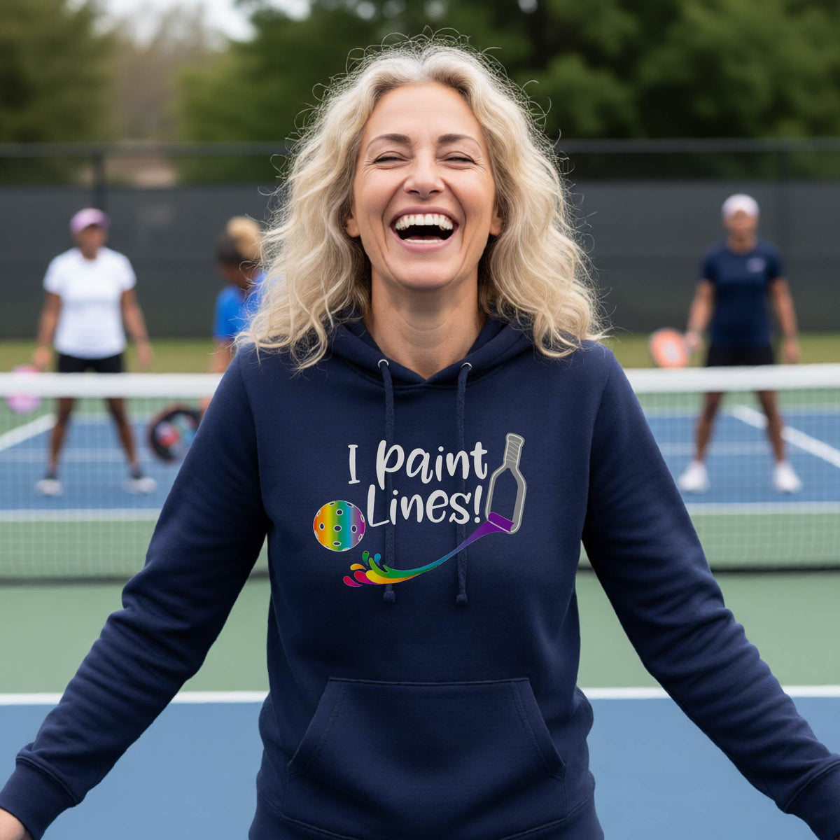 A woman wearing a navy blue fitted hoodie pullover with a graphic on the front that has white text "I PAINT LINES'  with a white pickleball paddle painting multi-colored lines leading a mutli-colored pickleball.