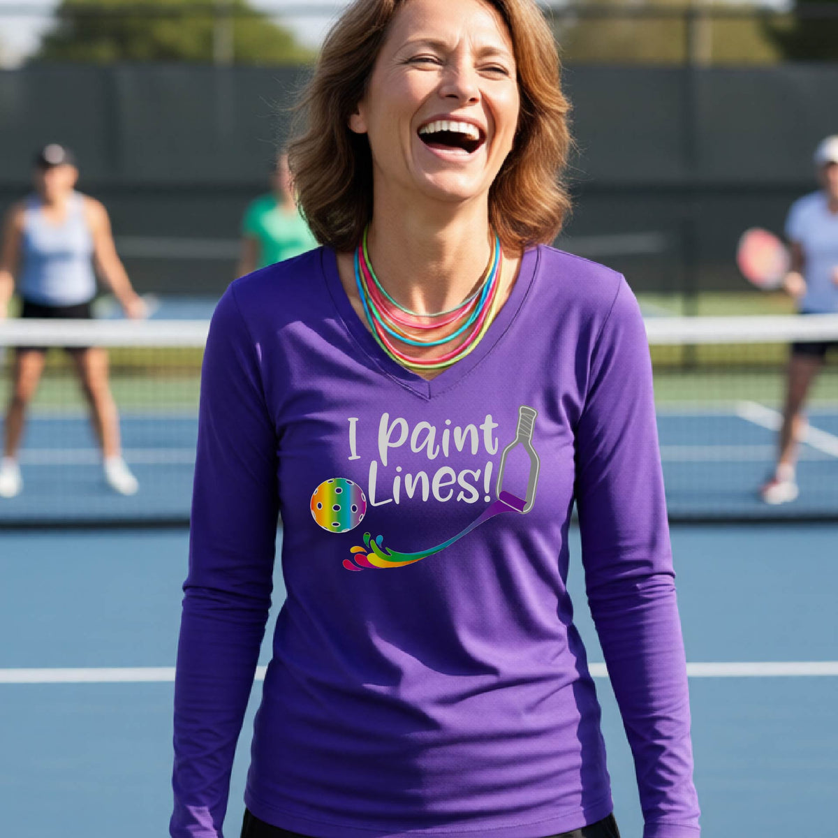 A close-up of a woman wearing a coral purple long-sleeve v-neck t-shirt with a graphic on the front that has white text "I PAINT LINES'  with a white pickleball paddle painting multi-colored lines leading a mutli-colored pickleball.