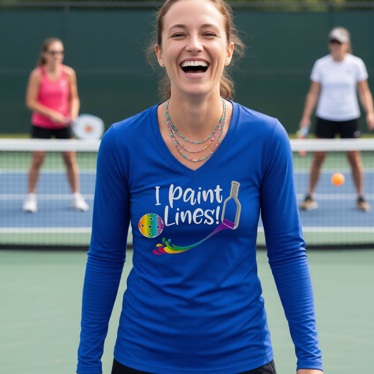 A close-up of a woman wearing a coral royal blue long-sleeve v-neck t-shirt with a graphic on the front that has white text "I PAINT LINES'  with a white pickleball paddle painting multi-colored lines leading a mutli-colored pickleball.