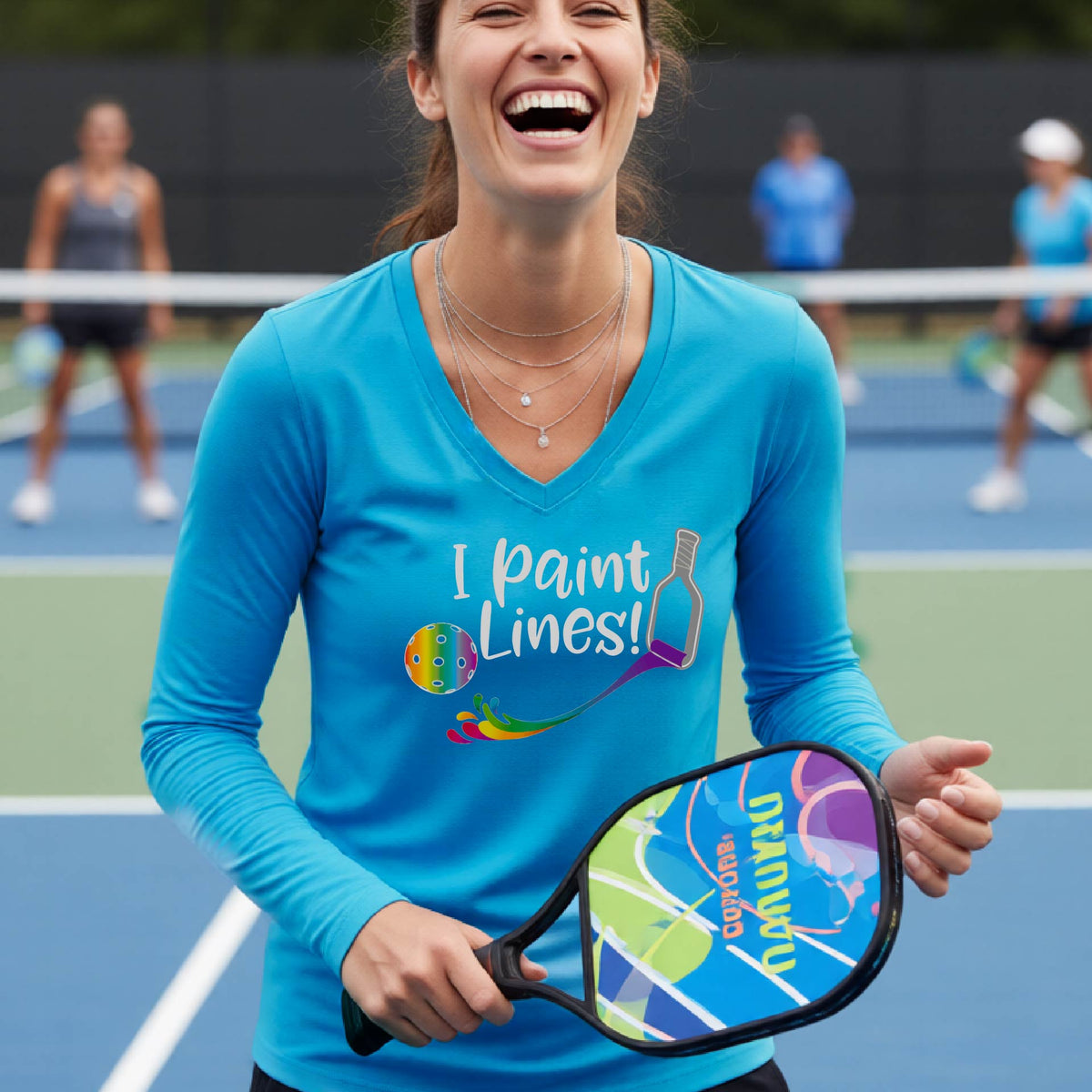 A close-up of a woman wearing a coral bright blue long-sleeve v-neck t-shirt with a graphic on the front that has white text "I PAINT LINES'  with a white pickleball paddle painting multi-colored lines leading a mutli-colored pickleball.